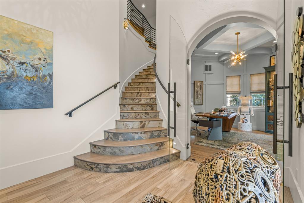 The curved marbled staircase and the first-floor office beyond a glass-and-iron arch: walnut waterfall desk, coffered ceiling, sputnik chandelier